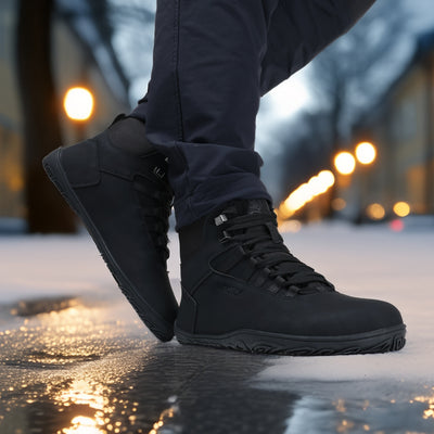 Käyttökuva noenmustista Uoma Talvi 3 -vaelluskengistä lumisella jalkakäytävällä, iltavalojen heijastuessa märästä pinnasta.