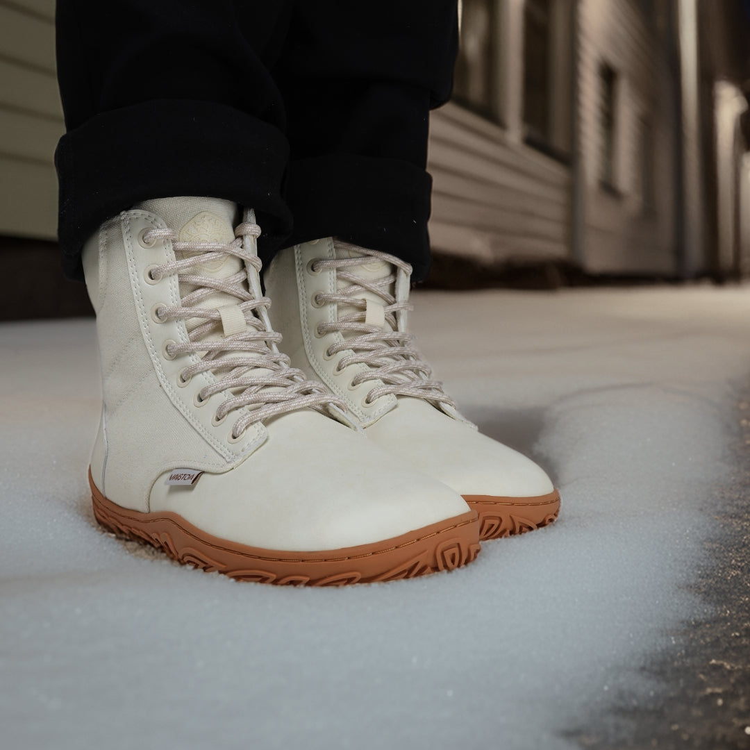 Luonnonvalkeat Vaistoa Hurmos 2 -paljasjalkamaiharit käytössä iltavalaistulla kadulla. Henkilö seisoo lumella rakennusten välissä, kengät korostuvat lämpimässä valossa.