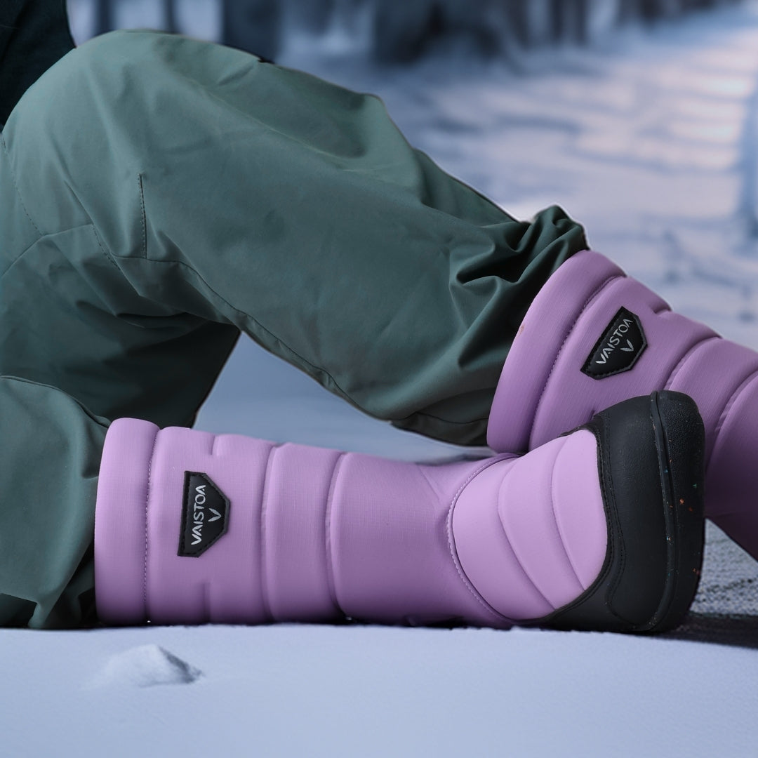 Vaistoa Kinos – lasten korkeavartiset paljasjalkakengät talveen, väri lila. Käyttökuva: edestä kuvattuna, lapsi istuu lumella siniset housut jalassa.