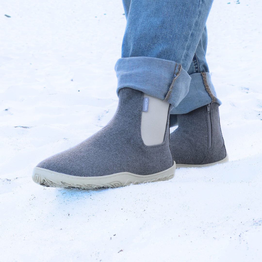Kuva henkilöstä sinisissä farkuissa, joka seisoo lumella helmenharmaat Vaistoa Sulo -huopanilkkurit jalassa. Kuvakulma etuviistosta korostaa kengän huopapinnan tekstuuria ja luonnollista askellusta tukevan pohjan rakennetta.